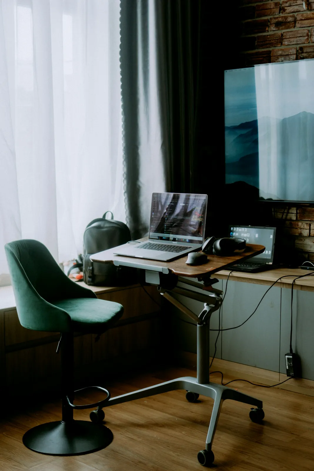 Laptop and monitor on a desk beside a window with a green office chair