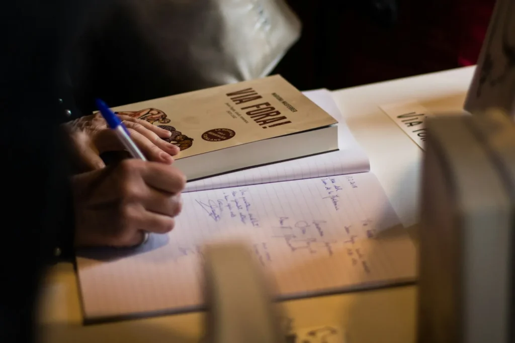 A person writing notes in a notebook beside an open book